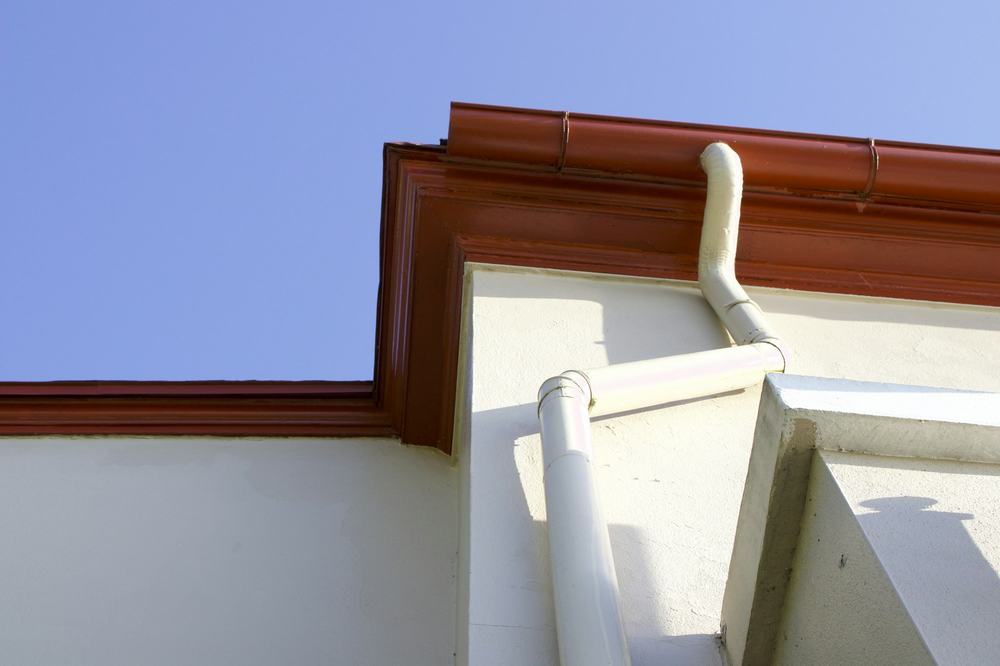 A gutter and downspout running along the roof edge of a white house against a blue sky