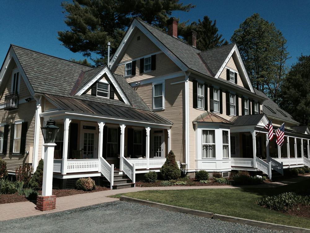 A gray New England house with white trim, gabled dormers and a wraparound front porch