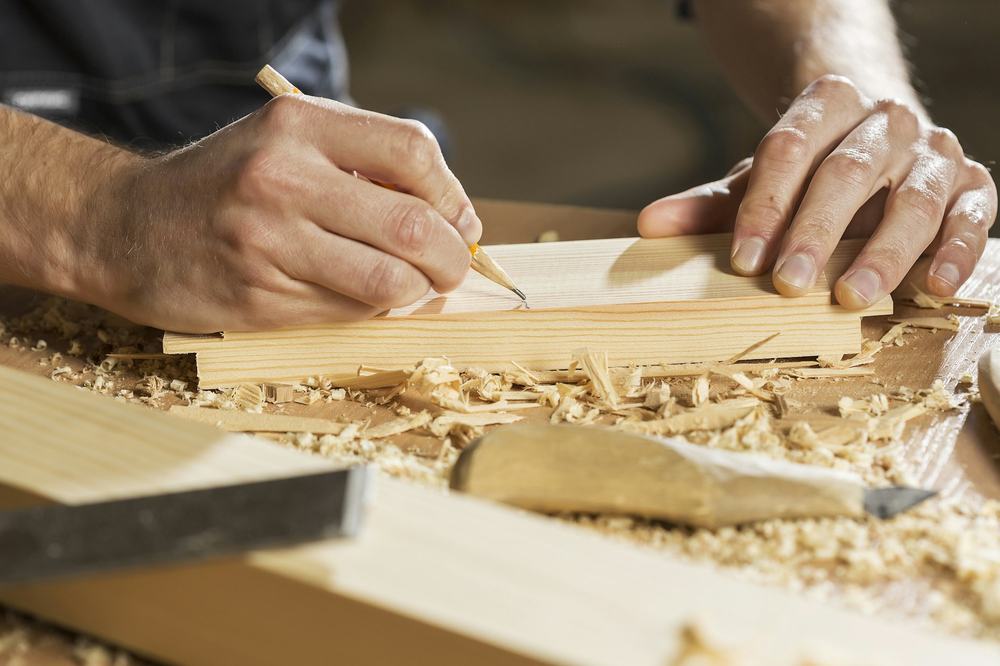 A carpenter's hands marking a cut line on a wooden board with a pencil, wood shavings on the bench